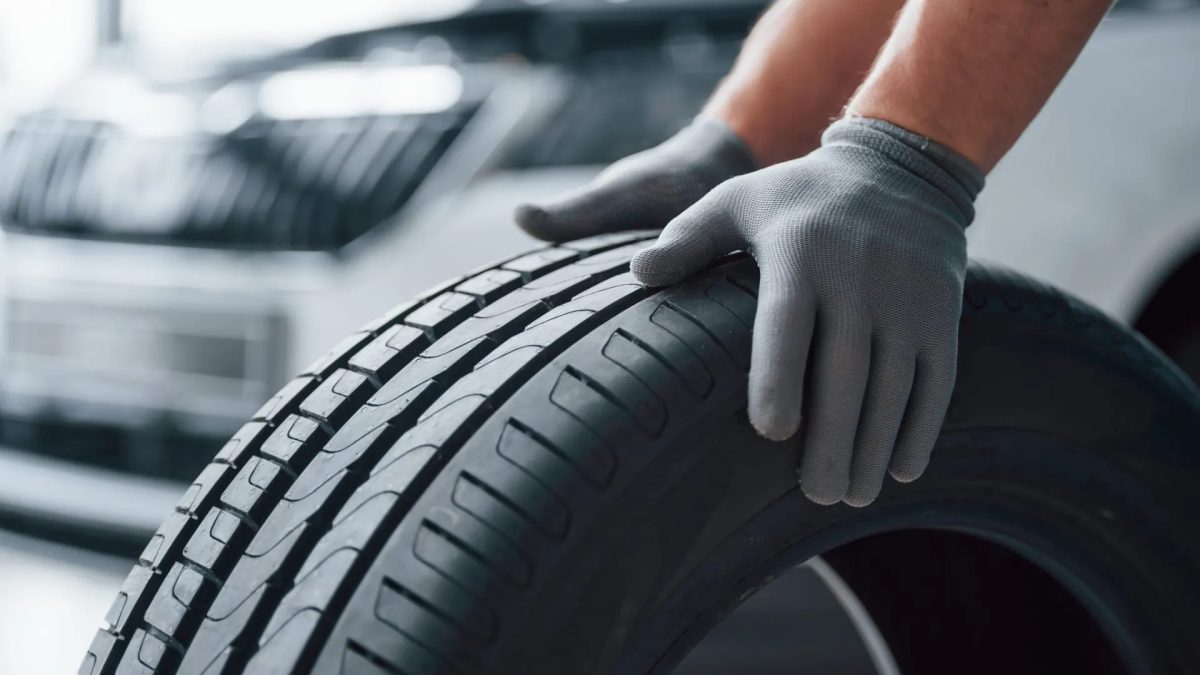 Mão com luva de proteção ajustando e inspecionando o pneu de um carro, com a banda de rodagem em destaque e o veículo ao fundo desfocado em ambiente urbano.