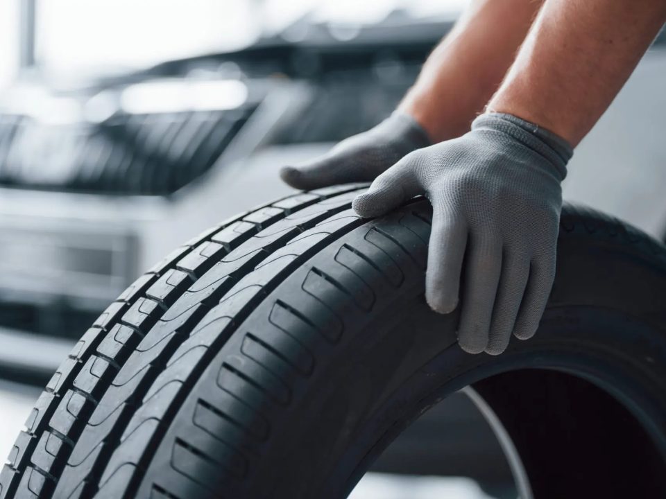 Mão com luva de proteção ajustando e inspecionando o pneu de um carro, com a banda de rodagem em destaque e o veículo ao fundo desfocado em ambiente urbano.