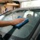 Pessoa passando pano de microfibra azul no para-brisa de um carro, limpando a superfície com gotas de água visíveis.