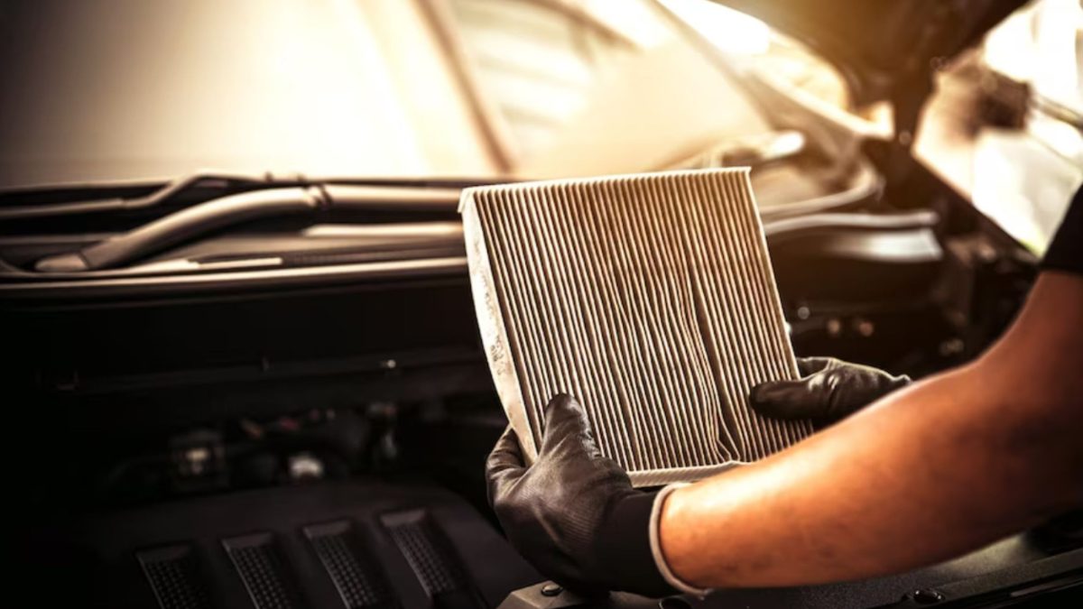 Mãos com luvas pretas segurando um filtro de cabine sujo, retirado de um carro com o capô aberto.
