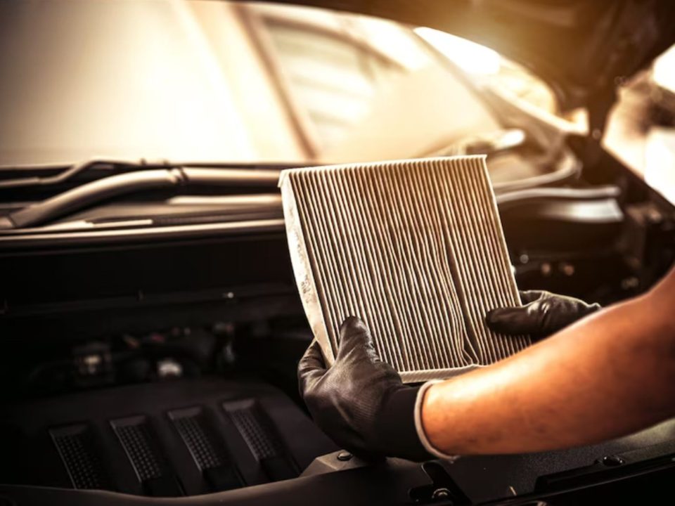 Mãos com luvas pretas segurando um filtro de cabine sujo, retirado de um carro com o capô aberto.
