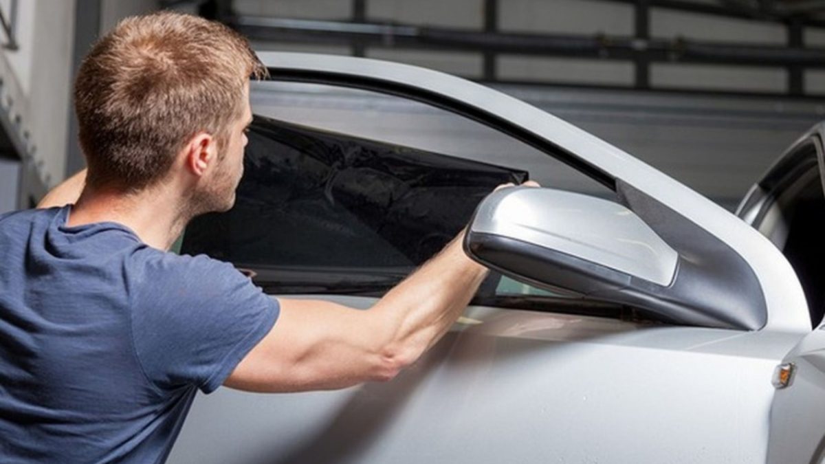 Homem aplicando película escura no vidro lateral dianteiro de um carro branco em uma garagem.