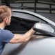 Homem aplicando película escura no vidro lateral dianteiro de um carro branco em uma garagem.