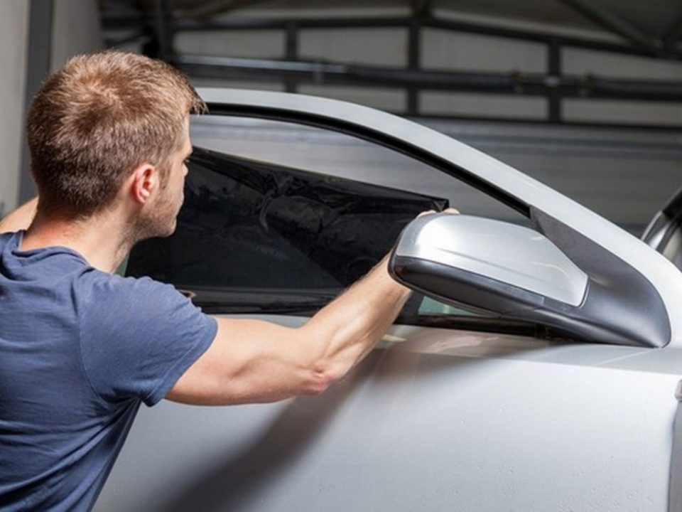 Homem aplicando película escura no vidro lateral dianteiro de um carro branco em uma garagem.