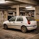 Fiat Palio branco parado em vaga de estacionamento coberto de prédio residencial, com poeira acumulada na pintura indicando longo período sem uso.