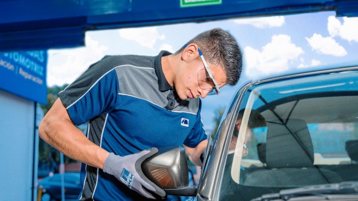 Técnico de uniforme azul e cinza instalando um retrovisor externo em um carro, usando luvas de proteção, em uma oficina automotiva.