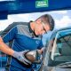 Técnico de uniforme azul e cinza instalando um retrovisor externo em um carro, usando luvas de proteção, em uma oficina automotiva.