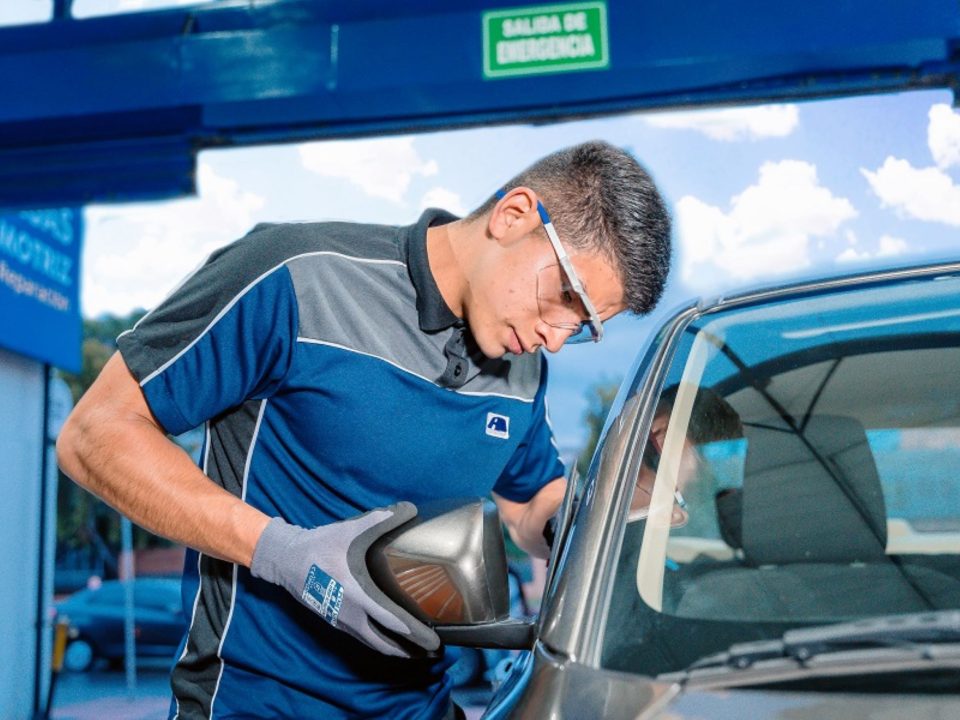 Técnico de uniforme azul e cinza instalando um retrovisor externo em um carro, usando luvas de proteção, em uma oficina automotiva.