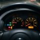 Painel de instrumentos de um carro com várias luzes de advertência acesas, visto através do volante, incluindo tacômetro e velocímetro iluminados.