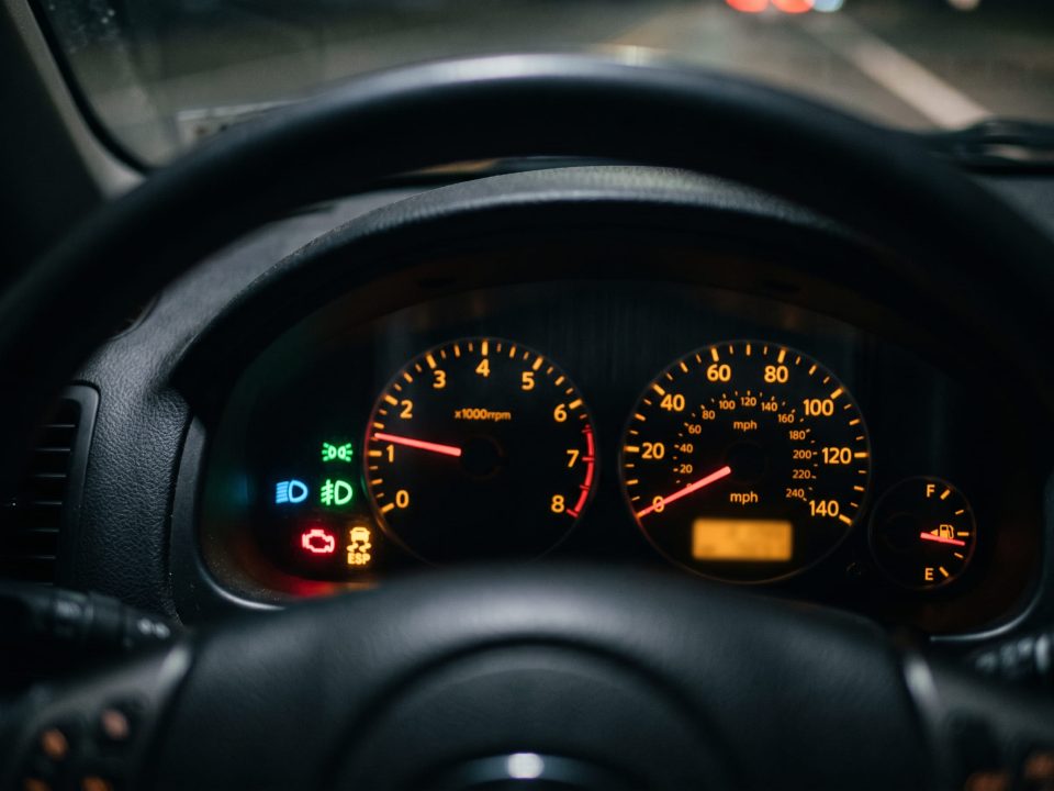 Painel de instrumentos de um carro com várias luzes de advertência acesas, visto através do volante, incluindo tacômetro e velocímetro iluminados.