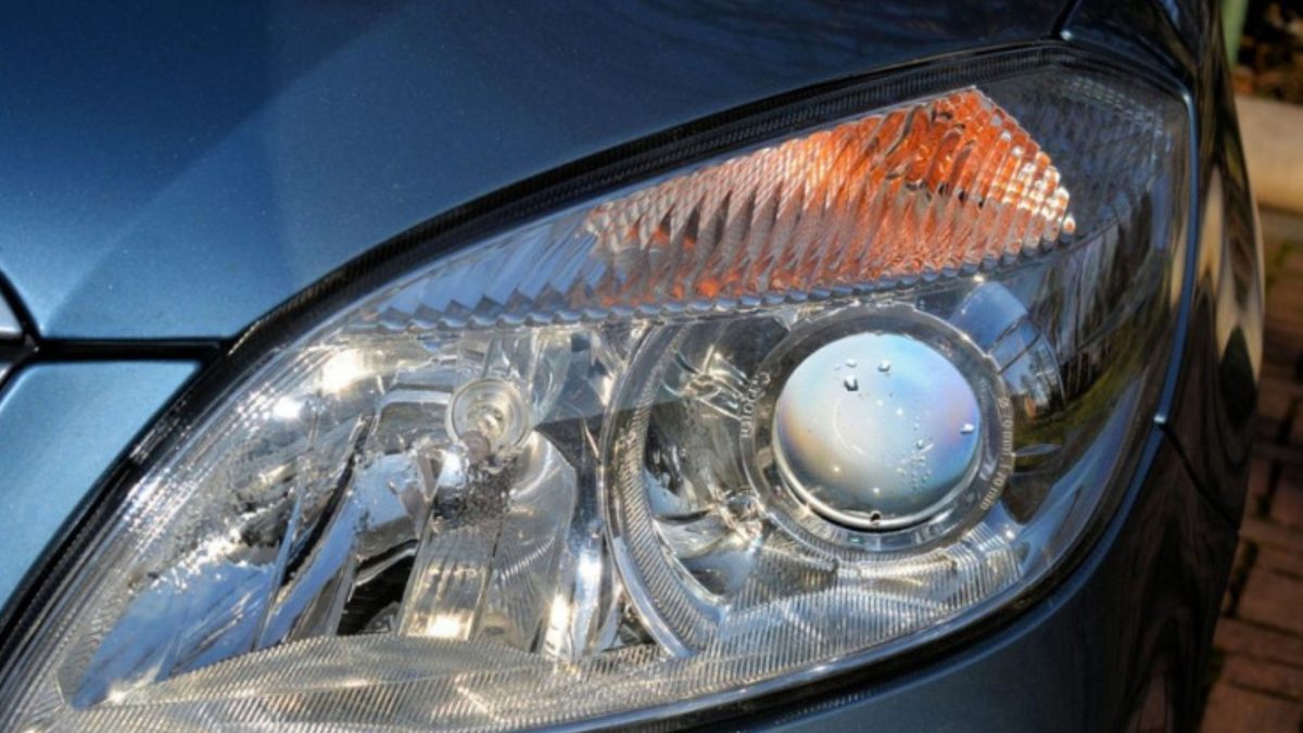 Farol dianteiro de um carro azul-acinzentado, com lente transparente, projetor redondo e luz de seta laranja.