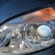 Farol dianteiro de um carro azul-acinzentado, com lente transparente, projetor redondo e luz de seta laranja.