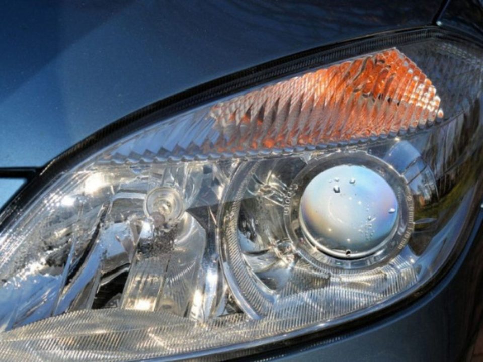 Farol dianteiro de um carro azul-acinzentado, com lente transparente, projetor redondo e luz de seta laranja.