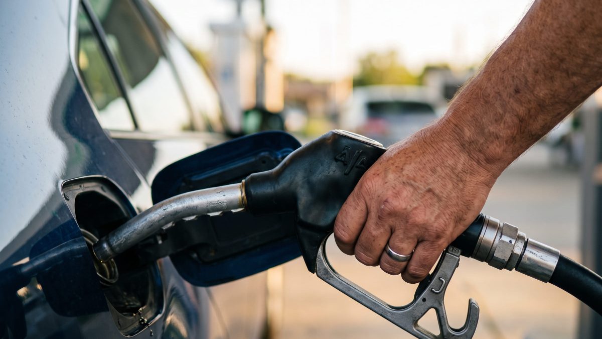 Foto de uma mão masculina segurando o bico da bomba de combustível durante o abastecimento de um carro em um posto de gasolina.