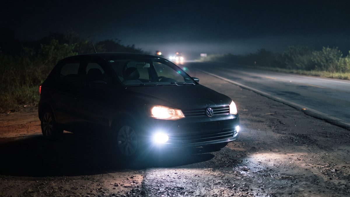Farol de milha aceso na dianteira de um Volkswagen Gol preto parado no acostamento de uma rodovia à noite.
