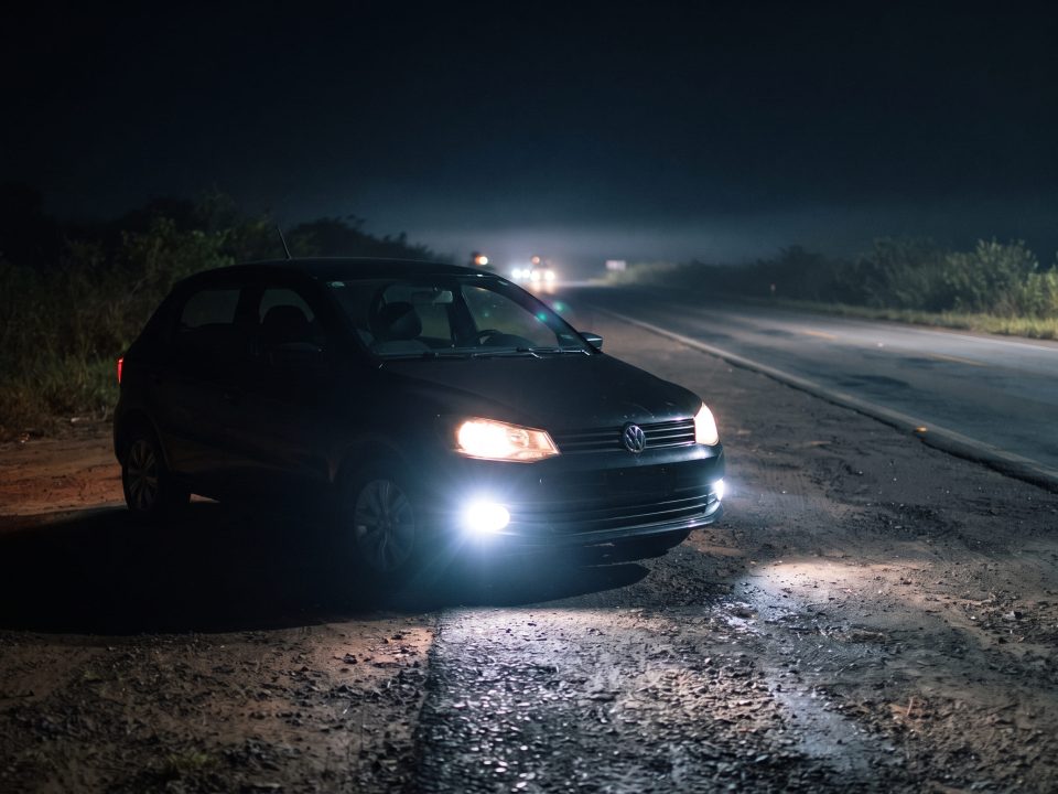 Farol de milha aceso na dianteira de um Volkswagen Gol preto parado no acostamento de uma rodovia à noite.