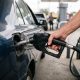 Mão segurando bico de combustível inserido no bocal de um carro azul em posto de gasolina.