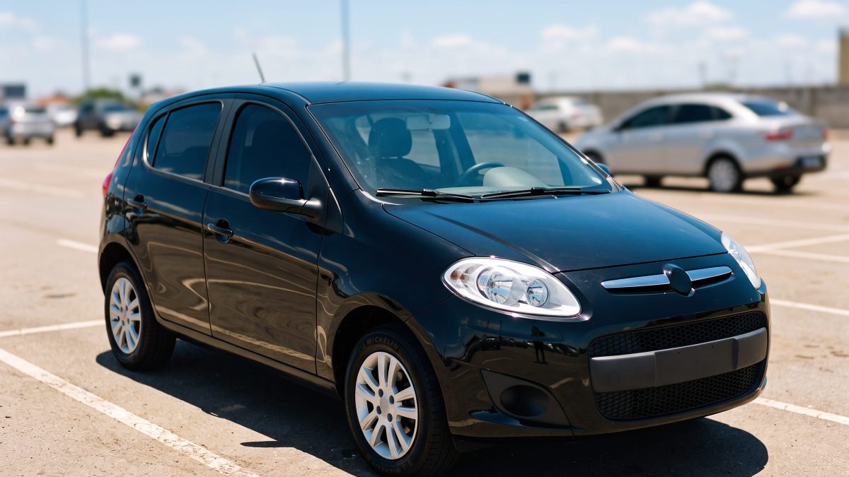 Fiat Palio preto estacionado sob sol forte em estacionamento aberto, com reflexo intenso na lataria.