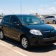 Fiat Palio preto estacionado sob sol forte em estacionamento aberto, com reflexo intenso na lataria.