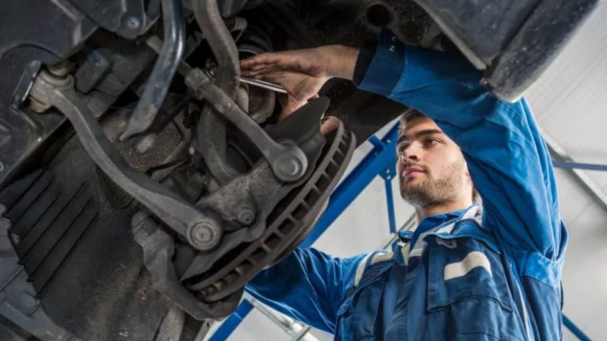 Mecânico em uma oficina usando luvas e trabalhando sob o veículo, com foco na área inferior do carro e na manutenção da suspensão.