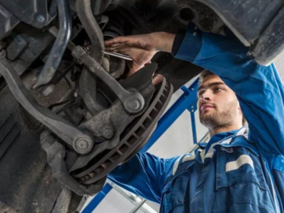 Mecânico em uma oficina usando luvas e trabalhando sob o veículo, com foco na área inferior do carro e na manutenção da suspensão.