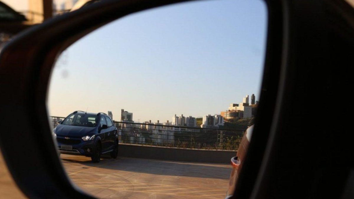Vista através do retrovisor externo de um carro, mostrando outro veículo estacionado em um estacionamento a céu aberto com prédios da cidade ao fundo.