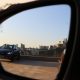 Vista através do retrovisor externo de um carro, mostrando outro veículo estacionado em um estacionamento a céu aberto com prédios da cidade ao fundo.