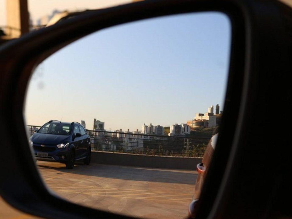 Vista através do retrovisor externo de um carro, mostrando outro veículo estacionado em um estacionamento a céu aberto com prédios da cidade ao fundo.