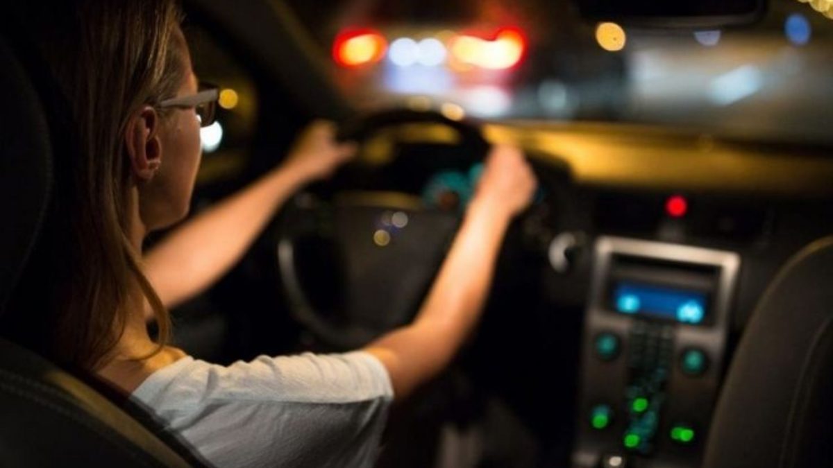 Motorista com as mãos no volante dirigindo à noite, com luzes desfocadas ao fundo e painel do carro iluminado.
