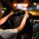 Motorista com as mãos no volante dirigindo à noite, com luzes desfocadas ao fundo e painel do carro iluminado.