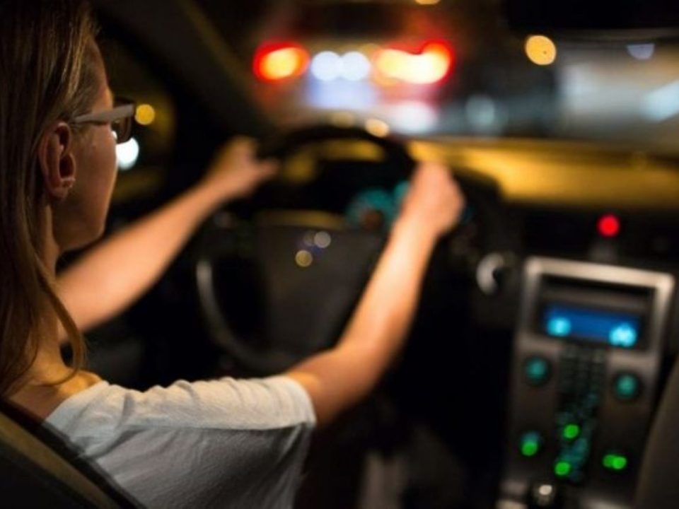 Motorista com as mãos no volante dirigindo à noite, com luzes desfocadas ao fundo e painel do carro iluminado.