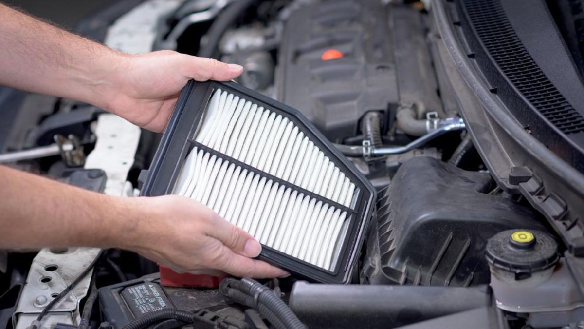 Mãos segurando um filtro de ar do motor sobre o compartimento aberto de um carro, mostrando as dobras do elemento filtrante.