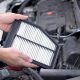 Mãos segurando um filtro de ar do motor sobre o compartimento aberto de um carro, mostrando as dobras do elemento filtrante.