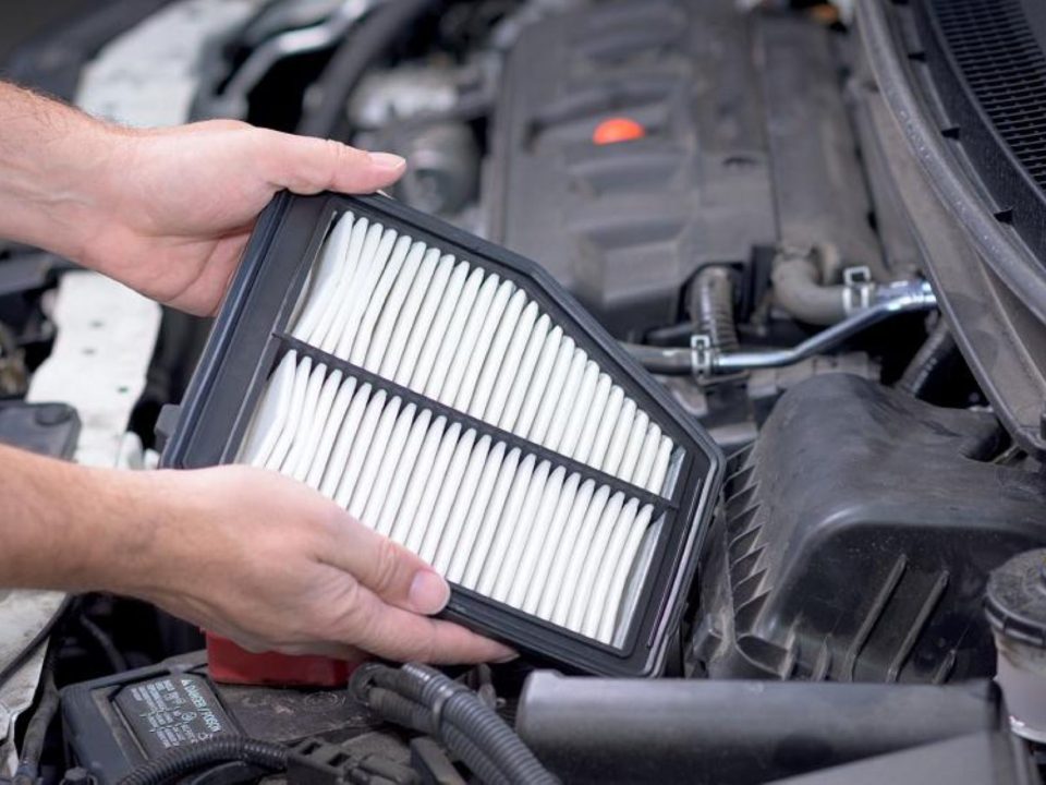 Mãos segurando um filtro de ar do motor sobre o compartimento aberto de um carro, mostrando as dobras do elemento filtrante.