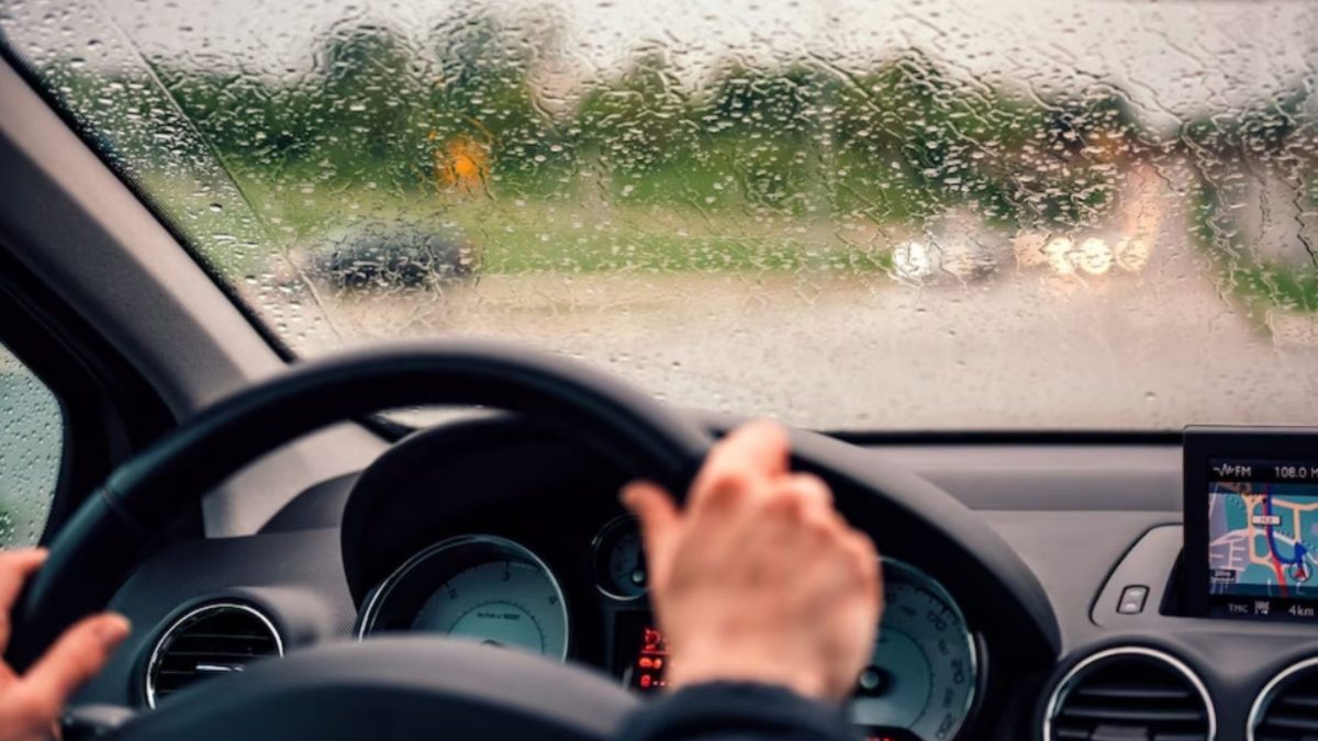 Vista interna de um carro em movimento na chuva, com a mão do motorista no volante e o para-brisa coberto de gotas de água, painel digital visível ao fundo.