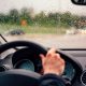 Vista interna de um carro em movimento na chuva, com a mão do motorista no volante e o para-brisa coberto de gotas de água, painel digital visível ao fundo.