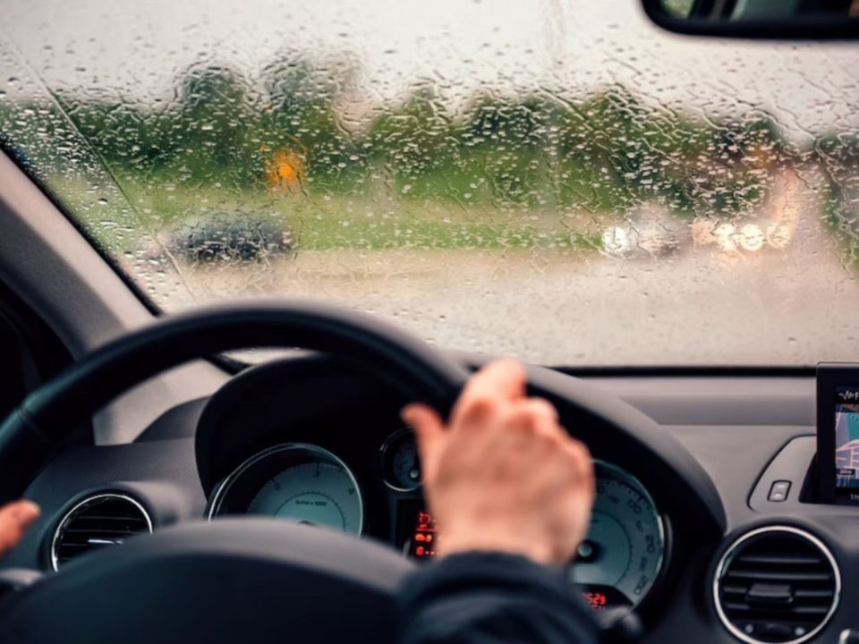 Vista interna de um carro em movimento na chuva, com a mão do motorista no volante e o para-brisa coberto de gotas de água, painel digital visível ao fundo.