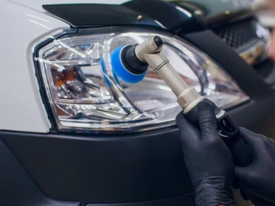 Profissional com luvas pretas usando politriz com boina azul para polir o farol de um carro branco.
