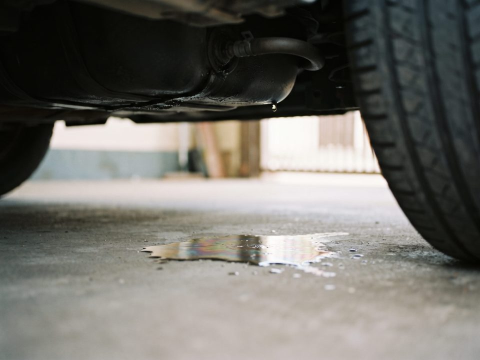 Mancha de gasolina no chão da garagem, embaixo da parte traseira de um carro, perto do tanque de combustível.