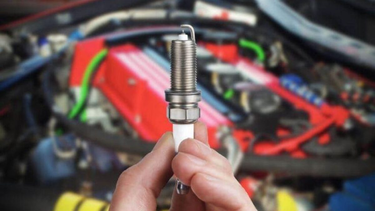 Mão segurando uma vela de ignição nova em frente ao motor aberto do carro.