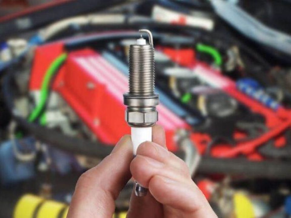 Mão segurando uma vela de ignição nova em frente ao motor aberto do carro.