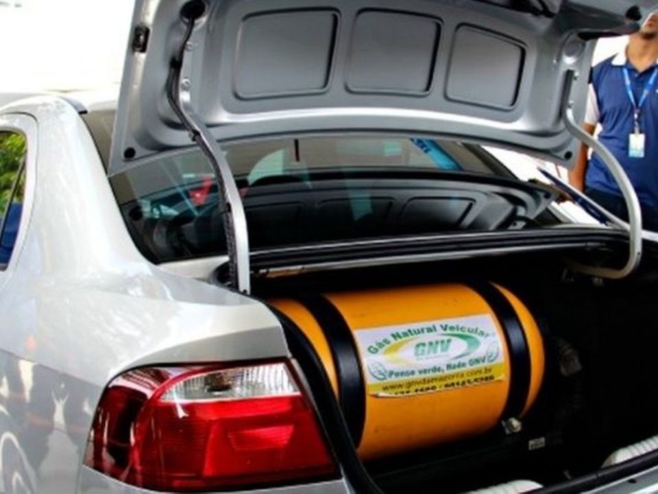 Porta-malas de carro branco aberto mostrando o cilindro amarelo de GNV instalado, com homem observando ao fundo.