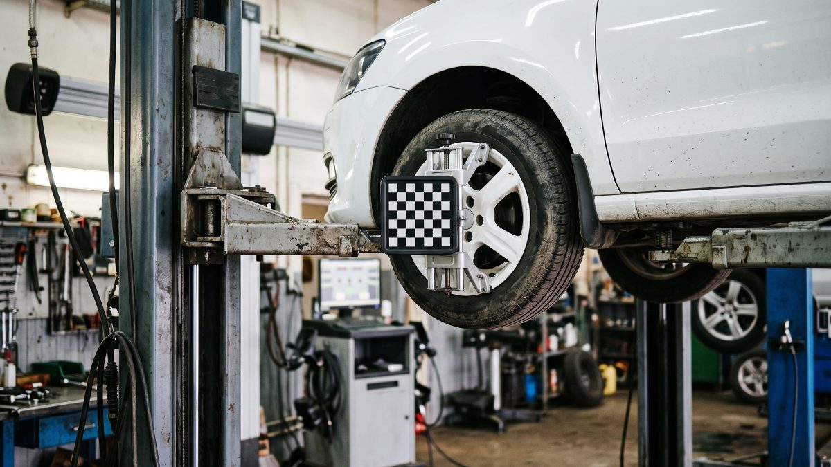 Carro branco elevado em oficina mecânica com braçadeira de alvo alinhador presa à roda dianteira, próximo ao equipamento de alinhamento de direção.