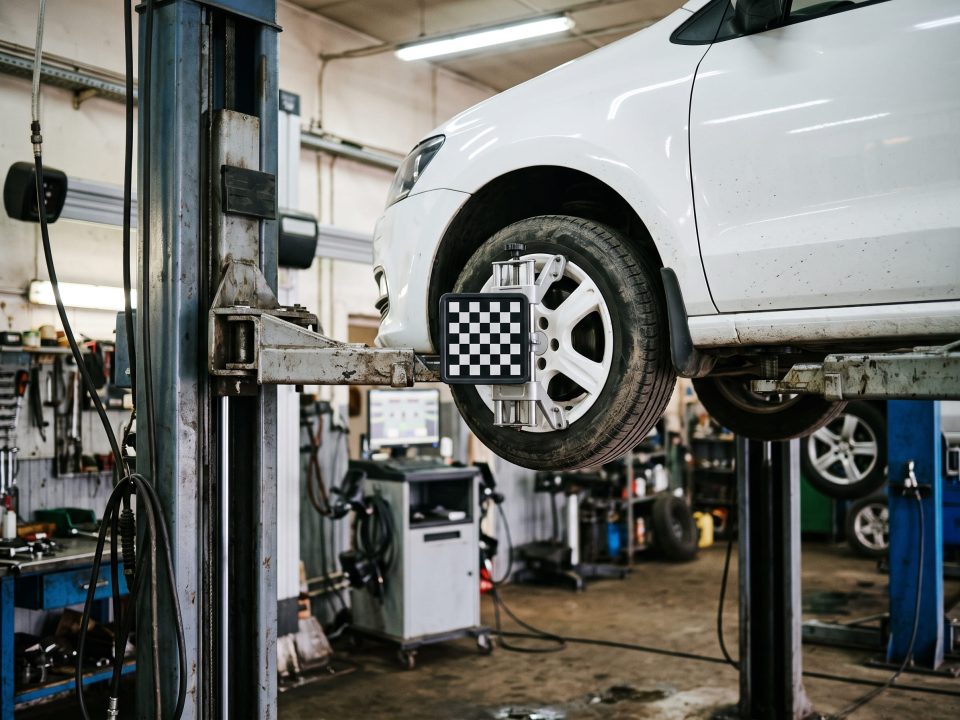 Carro branco elevado em oficina mecânica com braçadeira de alvo alinhador presa à roda dianteira, próximo ao equipamento de alinhamento de direção.