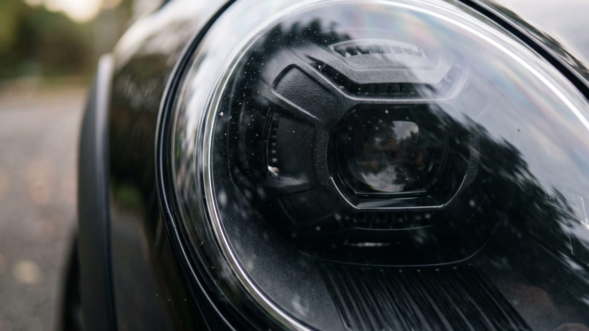 Imagem do farol dianteiro de um carro escuro, mostrando a máscara negra ao redor da lente central e o acabamento cromado na borda externa do conjunto óptico.