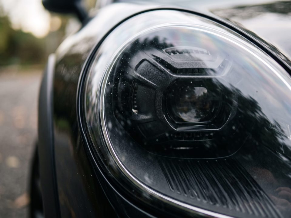 Imagem do farol dianteiro de um carro escuro, mostrando a máscara negra ao redor da lente central e o acabamento cromado na borda externa do conjunto óptico.