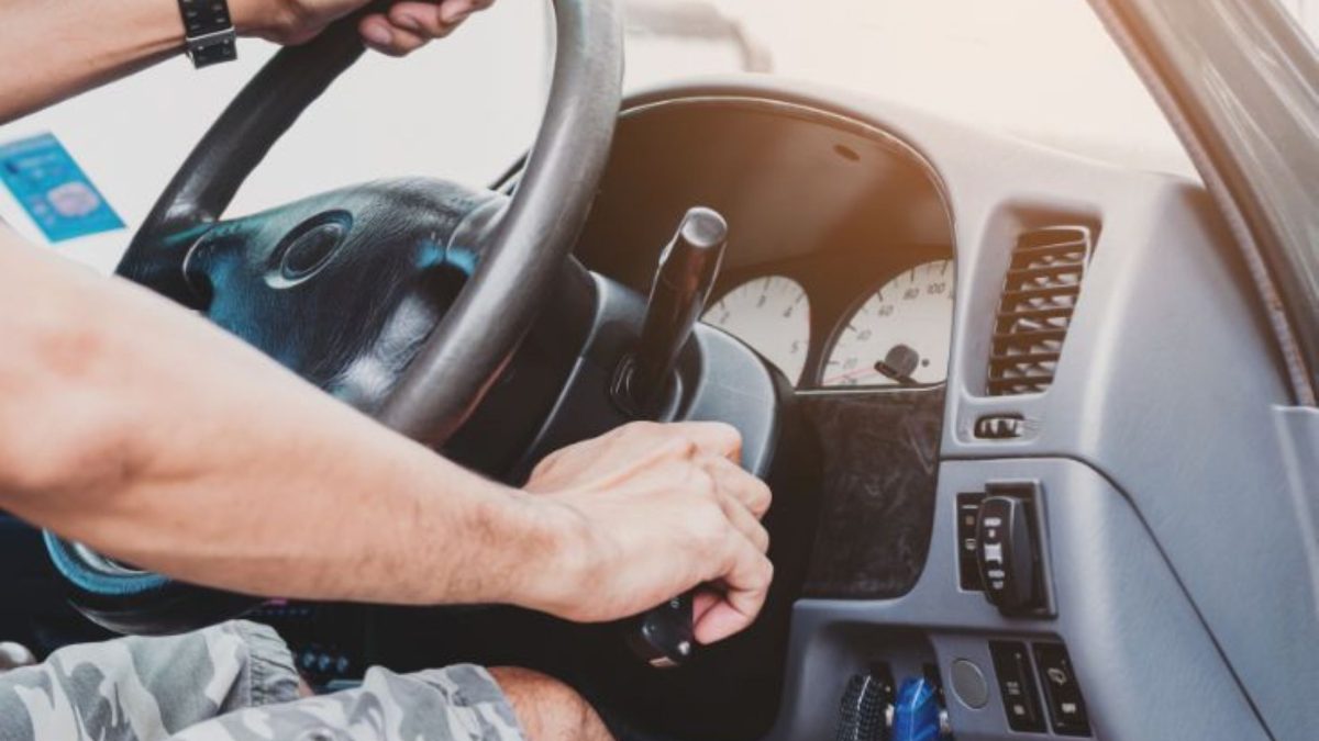 Mão de motorista girando a chave na ignição de um carro, com o volante e o painel de instrumentos ao fundo.