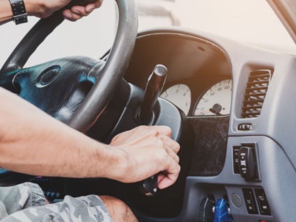 Mão de motorista girando a chave na ignição de um carro, com o volante e o painel de instrumentos ao fundo.