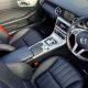 Interior de carro esportivo com bancos de couro preto, costura vermelha contrastante, cinto de segurança vermelho e volante multifuncional com câmbio automático ao centro.