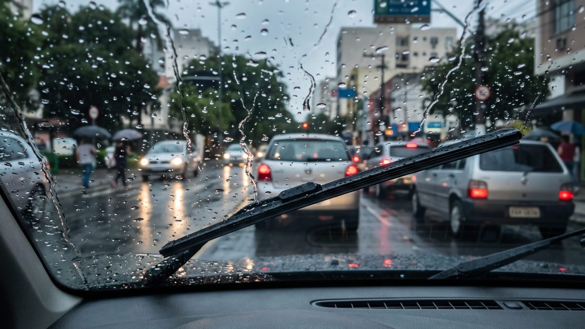 Foto feita de dentro do carro com palheta do limpador de para-brisa em funcionamento durante chuva intensa em via urbana.
